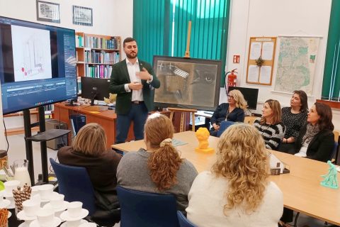 Grupa osób siedzi przy stole w pokoju. Na przodzie stoi mężczyzna przy ekranie.