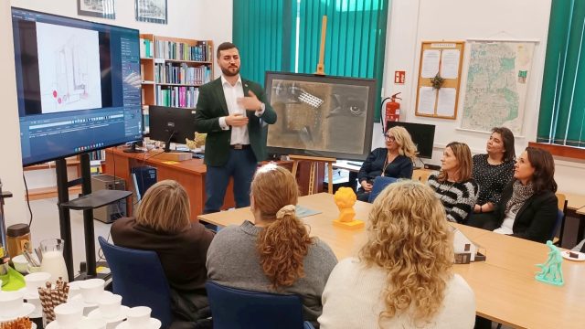 Grupa osób siedzi przy stole w pokoju. Na przodzie stoi mężczyzna przy ekranie.