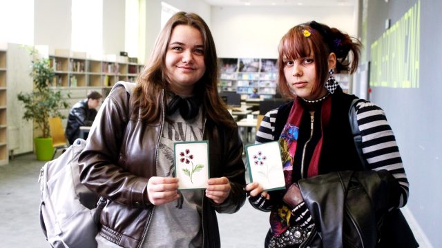 Dwie osoby stoją w bibliotecznej przestrzeni, trzymając w rękach kartki z rysunkami kwiatów