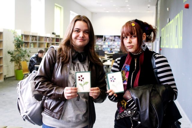 Dwie osoby stoją w bibliotecznej przestrzeni, trzymając w rękach kartki z rysunkami kwiatów