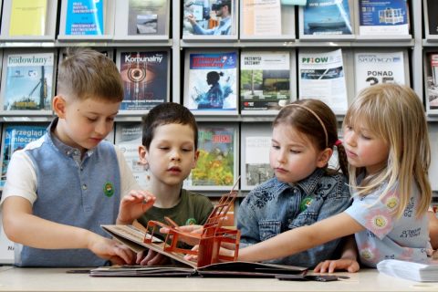 Grupa dzieci ogląda materiały edukacyjne przy stole w otoczeniu półek z publikacjami
