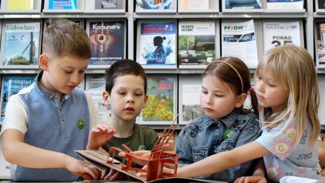 Grupa dzieci ogląda materiały edukacyjne przy stole w otoczeniu półek z publikacjami