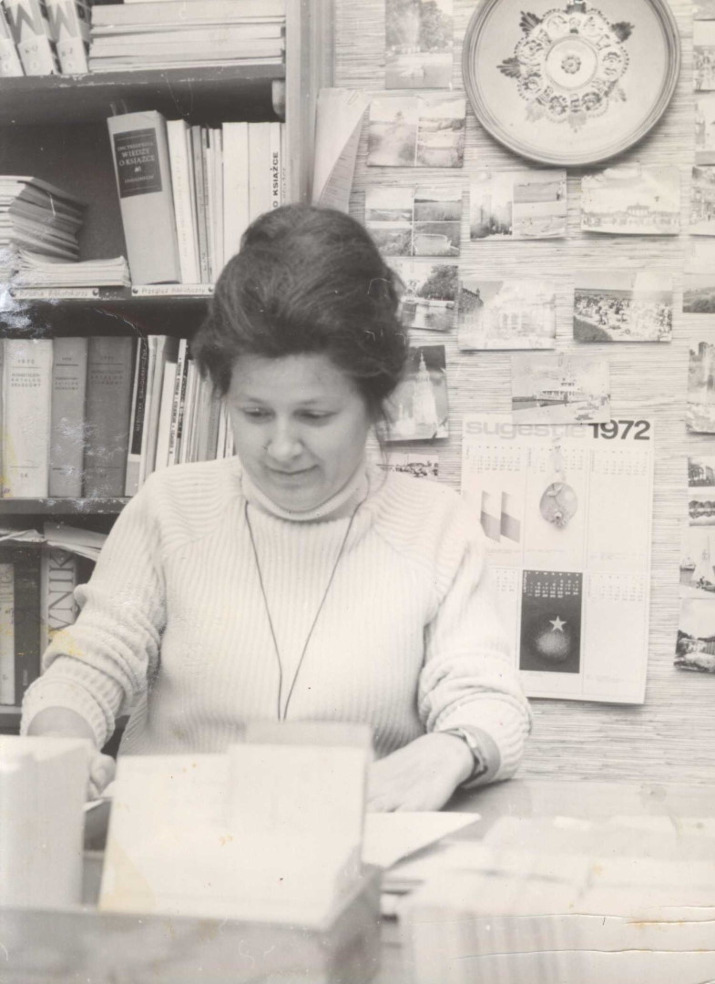 Przy biurku zapełnionym dokumentami siedzi kobieta, Dyrektor Biblioteki Politechniki Białostockiej mgr Regina Szymańska. Za jej plecami stoi regał z książkami.