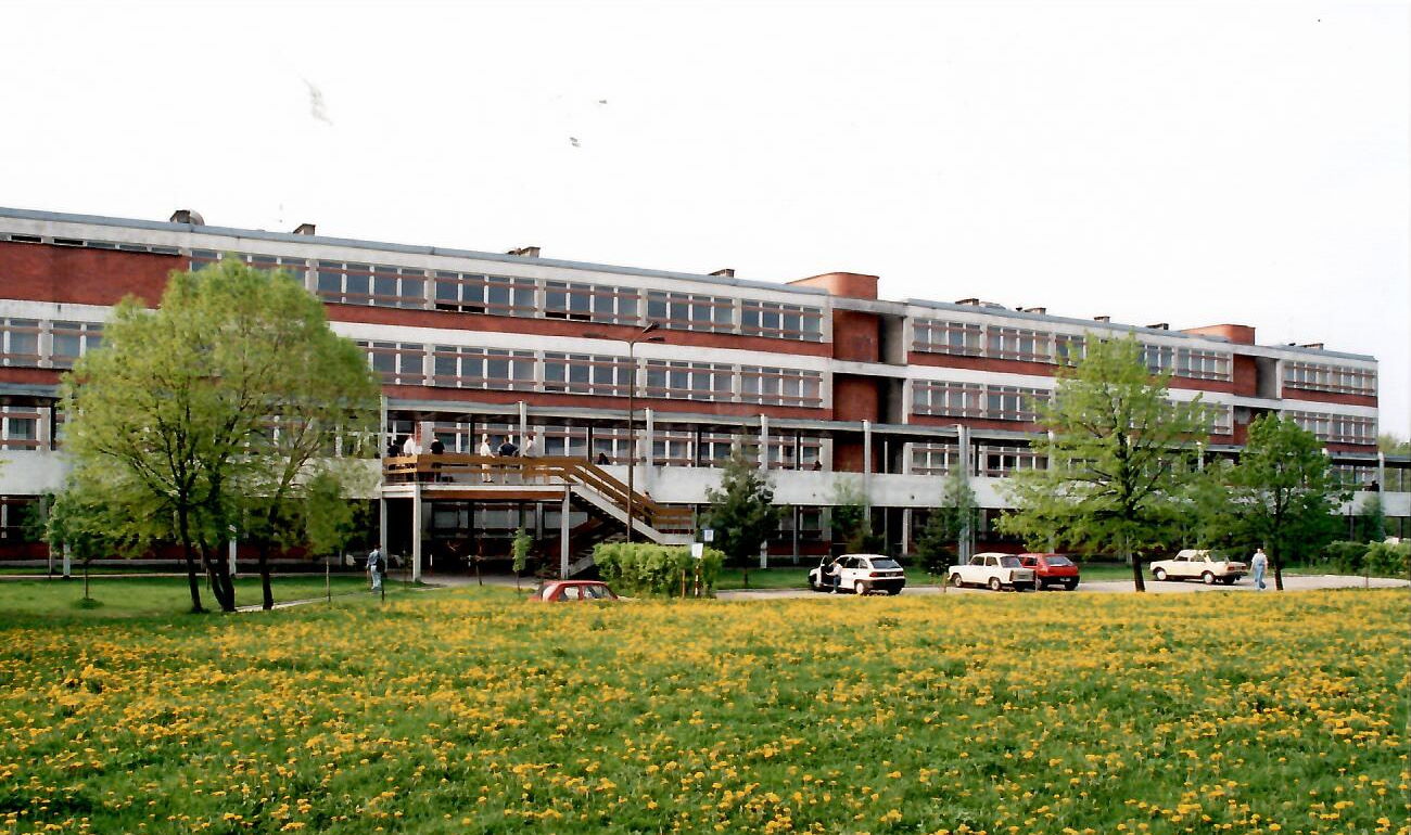 Widok jednego z budynków na kampusie Politechniki Białostockiej. Przed obiektem parking z nielicznymi samochodami, zielone drzewa i kwitnąca na żółto łąka.