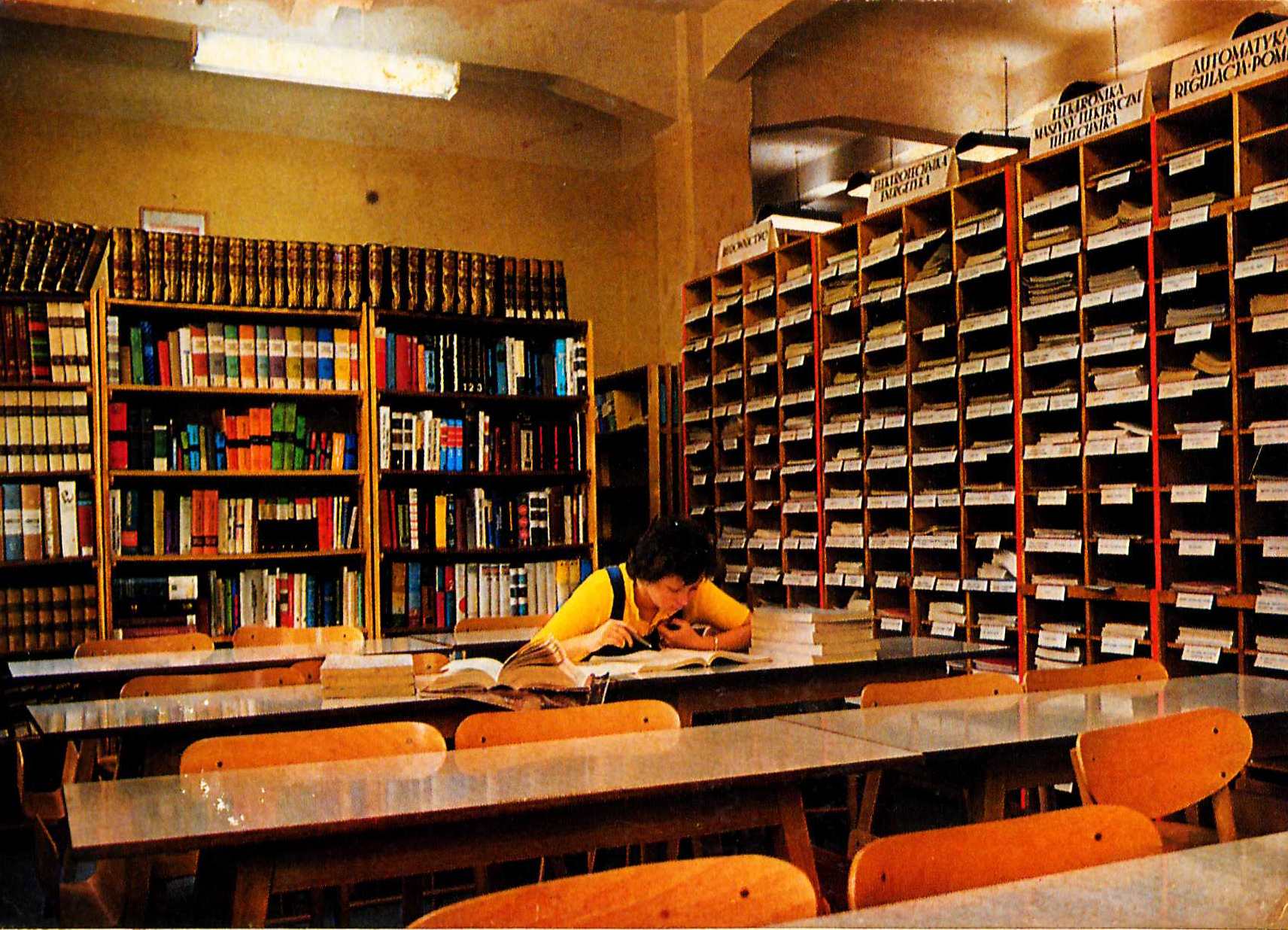 Pomieszczenie z dwoma ścianami regałów książek. Stojące w rzędach stoły z krzesłami, przy jednym z nich siedzi pochylona nad książkami kobieta.