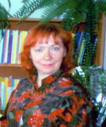 Portret rudowłosej kobiety, dyrektor Biblioteki Politechniki Białostockiej, mgr Heleny Kotkowskiej, patrzącej wprost, siedzącej na tle regału z książkami i paprotki.