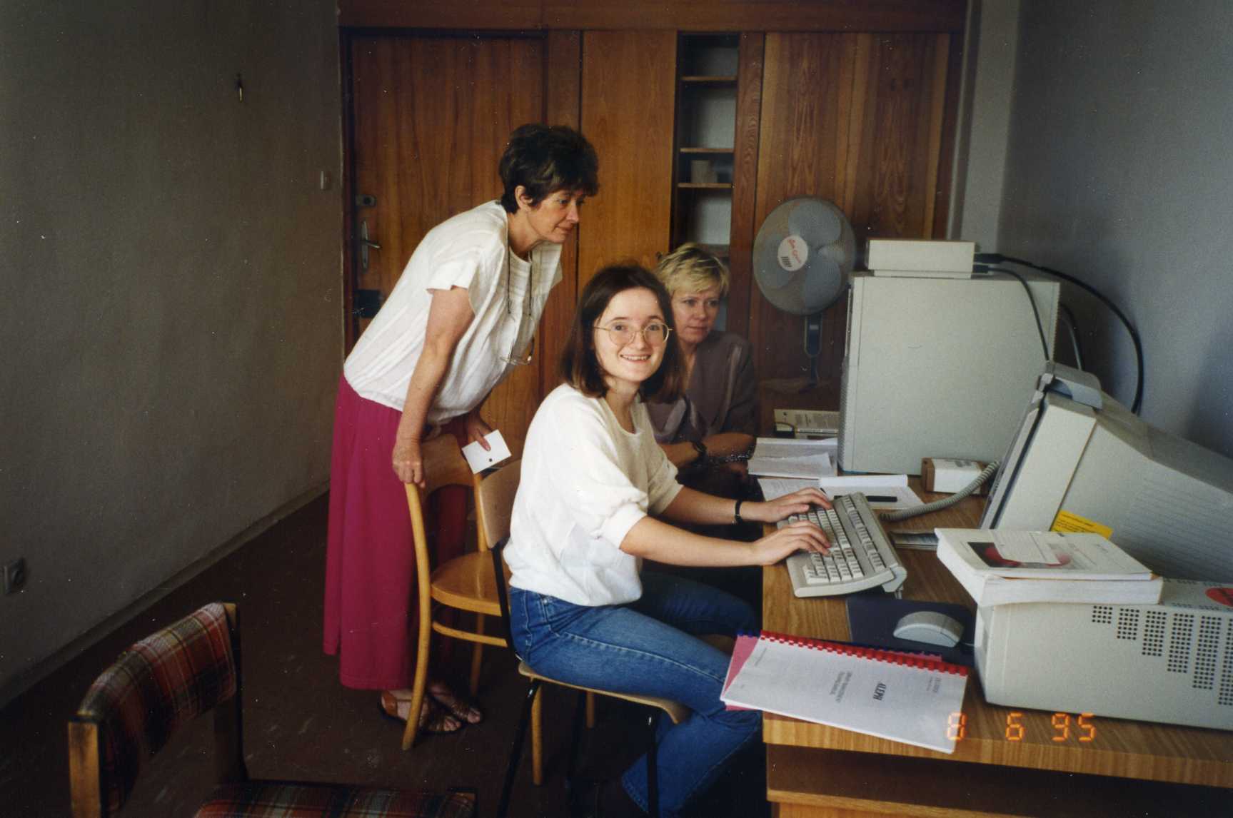 Pomieszczenie biurowe, pod lewą ścianą stoi biurko ze sprzętem komputerowym, dwie kobiety siedzą na krzesłach, a jedna stoi opierając się o poręcz krzesła, z tyłu na ścianie szafa i drzwi.