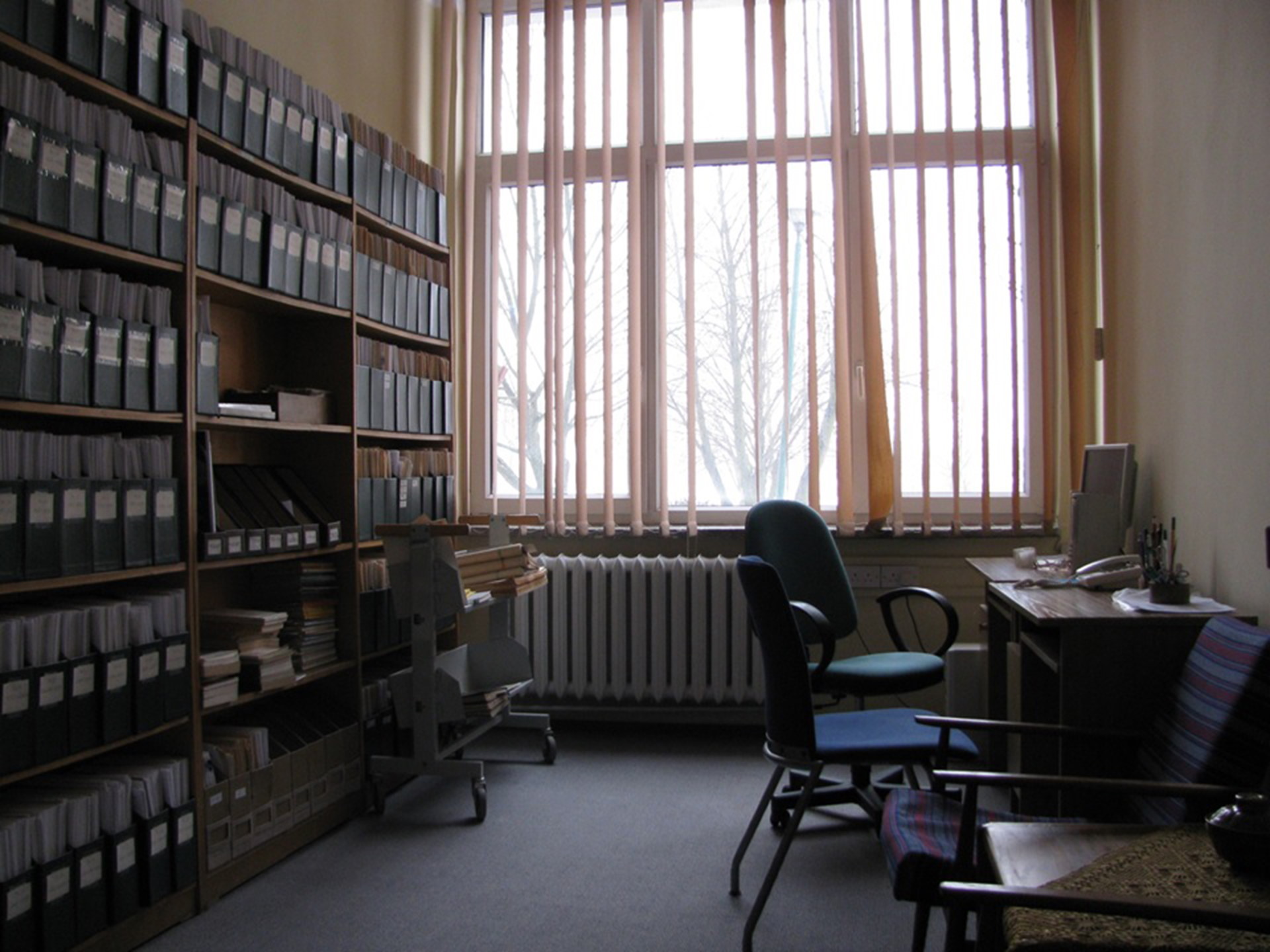 Pomieszczenie biurowe, w którym po prawej stoi biurko i dwa krzesła natomiast po lewej regał pełen segregatorów w jednolitym ciemnym kolorze, na środkowej ścianie okno przesłonięte pionowymi żaluzjami.