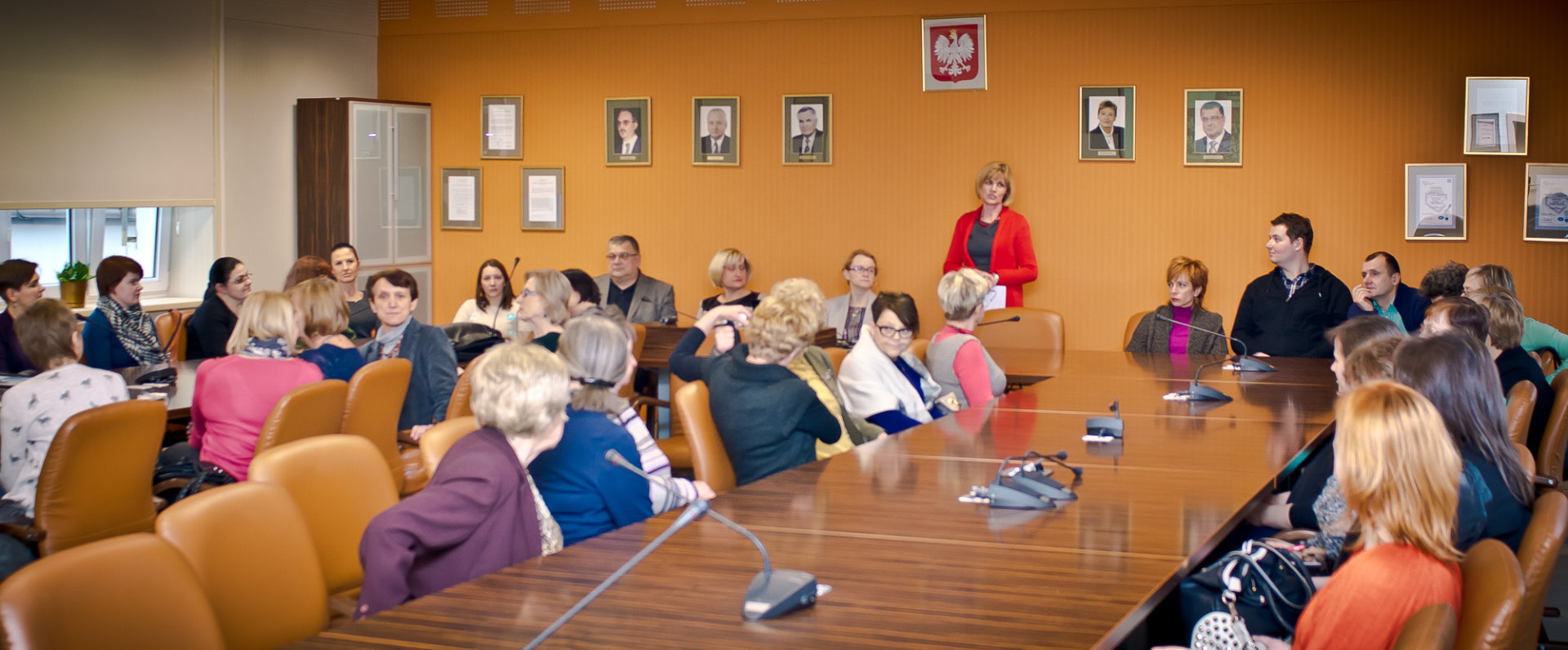Grupa ludzi siedząca przy stołach ustawionych w kształt podkowy, na stołach stoją mikrofony, w tle ściana udekorowana godłem Polski oraz portretami ludzi.