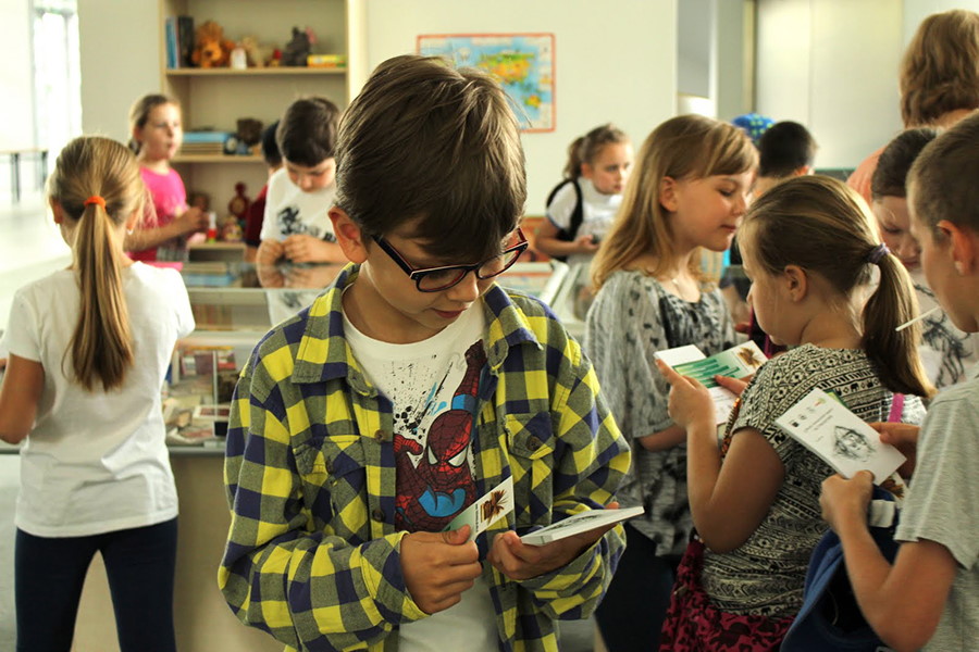 Chłopiec w okularach trzymający zakładkę i książkę, w tle grupa dzieci z książkami w dłoniach.