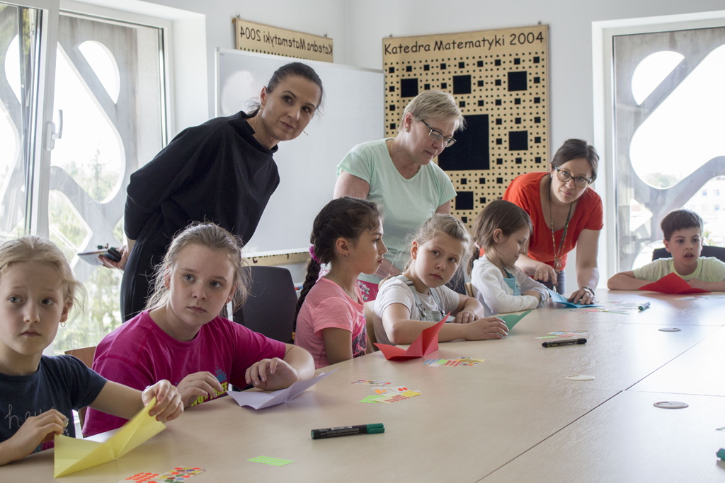 Sześcioro dzieci i trzy kobiety uczestniczą w zajęciach plastycznych. Przed dziećmi ułożone kolorowe kartki papieru i mazaki.