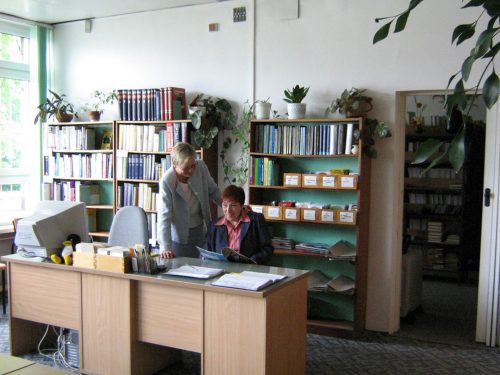 Pokój z podwójnym biurkiem i regałami ustawionymi wzdłuż ściany. Za biurkiem dwie rozmawiające kobiety. Na biurku ustawiony komputer. Po lewej stronie duże okno.