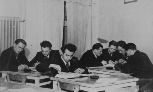 Zdjęcie czarno-białe. Siedmiu mężczyzn siedzi przy trzech kwadratowych stolikach. Zajmują się czytaniem książek i wspólną nauką. Na stolikach widać popielniczki.