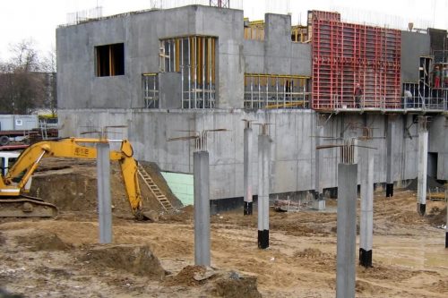 Za sześcioma wysokimi betonowymi słupami, stoi żółta koparka. Po prawej stronie duży betonowy budynek w stanie surowym. Na rusztowaniach zaczepionych na bryle budynku pracują robotnicy.