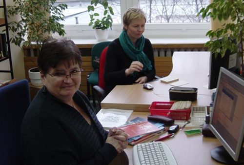 Dwie kobiety o krótkich włosach, w tym jedna w okularach, siedzą przy biurkach przed monitorami. Na blatach leżą książki, koszyczki z kartkami, długopis, etui. W tle widoczne są duże kwiaty. 