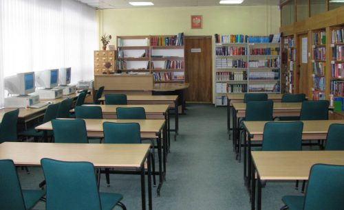 Pomieszczenie czytelni. W dwóch rzędach ustawione są biurka z zielonymi krzesłami. Z lewej strony wzdłuż okien stoją biurka z komputerami i monitorami. W tle duże biurko z nadstawką, za którym znajdują się regały z książkami. Po środku są brązowe drzwi, nad którymi wisi godło.