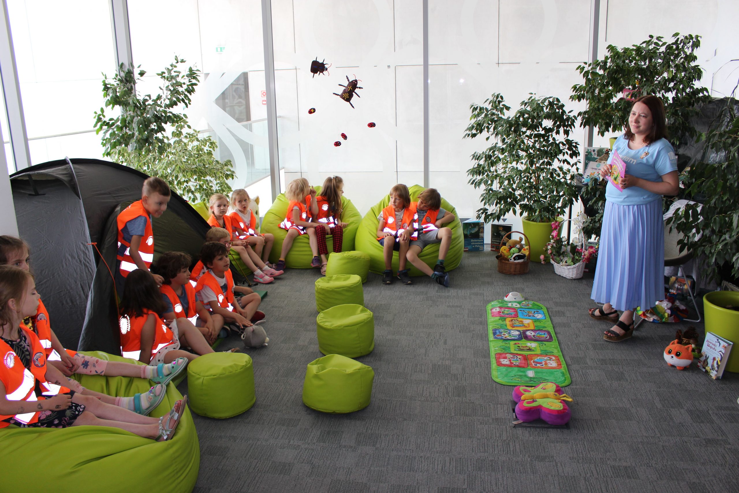 Przedszkolaki siedzą na zielonych pufach i słuchają czytanej książki przez bibliotekarza.