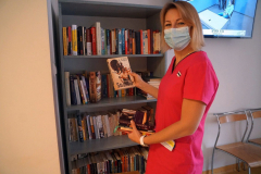 Uśmiechnięta kobieta w masce prezentująca książki na tle regału.