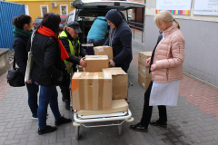 Przed wejściem do szpitala grupa osób wypakowuje z samochodu kartony z książkami.