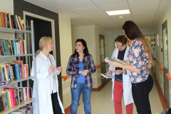 Opis alternatywny: Trzy kobiety i jeden mężczyzna rozmawiają w korytarzu szpitala przed regałem z książkami.