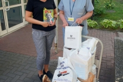 Dwie uśmiechnięte kobiety w okularach przed budynkiem Specjalistycznego Szpitala Onkologicznego. Przed  nimi platforma z torbami.