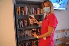 Uśmiechnięta kobieta w masce prezentująca książki na tle regału.