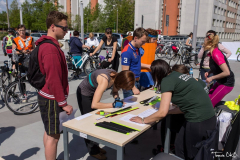 Grupa osób rejestrujących się na rajd rowerowy. Na stole leżą odblaskowe opaski i kartki papieru. W tle widać rowerzystów z rowerami.
