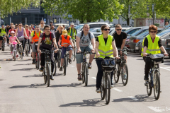 Grupa uśmiechniętych rowerzystów jedzie ulicą. Część z nich jest w odblaskowych kamizelkach.