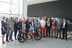 Grupa organizatorów i ambasadorów kampanii "Sam zadbaj o swoje bezpieczeństwo" stoi w szeregu. Przed nimi znajduje się czarno-czerwony rower.