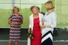 Od lewej widać uśmiechniętą kobietę z czerwoną różą w ręku, drugą trzymającą czerwoną różę i okrągłe pudełko, która rozmawia z dyrektor Marią Czyżewską.