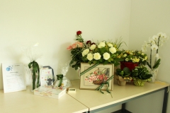 Stół zastawiony prezentami. Znajdują się na nim certyfikat w ramce, laurka z misiem, czekoladki w pudełku,  filiżanka, kwiaty i obraz w ramce przedstawiający kwiaty.
