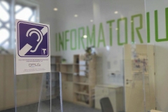 Symbol pętli indukcyjnej. Stanowisko obsługi czytelnika w Oddziale Informacji Naukowej. W tle regały z książkami i duży napis na ścianie Informatorium.