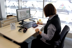 Stanowisko komputerowe dla osób z niepełnosprawnościami. Na biurku znajdują się: monitor, lampka, klawiatura Magic Keyboard, mysz Big Track oraz słuchawki. Przy biurku siedzi użytkowniczka, korzystająca z lupy elektronicznej.