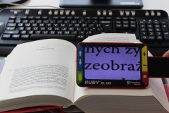 Otwarta książka. Na niej leży lupa elektroniczna Ruby XL HD powiększająca tekst w książce.