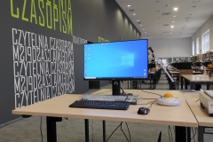 Stanowisko komputerowe dla osób z niepełnosprawnościami . Na biurku znajdują się: monitor, klawiatura, klawiatura Magic Keyboard, mysz Big Track,  słuchawki oraz lupa elektroniczna Ruby XL HD.  W tle stanowiska komputerowe dla czytelników.