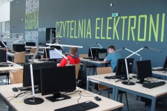 Dwóch studentów siedzących za komputerami w  Czytelni Elektronicznej. W tle zielony napis na ścianie.
