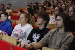 Grupa osób siedząca na krzesłach stacjonarnych. W pierwszym rzędzie młoda kobieta i trzech młodych mężczyzn.