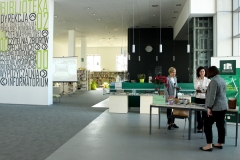 Duża przestrzeń biblioteczna.  Po prawej stronie wokół stołu zapełnionego przedmiotami stoją trzy kobiety. Po lewej stronie wysoka, jasna ściana z napisami. W oddali regały z książkami.