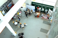 Widok z góry na hol, na którym stoją stoliki z krzesełkami. Osoby oglądają kolorowe stanowiska stojące przy ścianie.