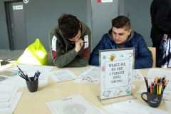 Na stole rozłożone karty oraz kubki z kolorowymi kredkami. Dwaj chłopcy pochyleni nad arkuszami.