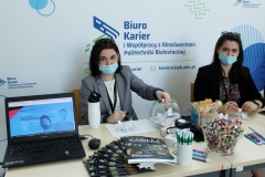 Kobiety w maseczkach na twarzy trzymają w ręce małe, złożone karteczki. Przed nimi stoi laptop i gadżety promocyjne.