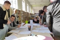 Grupa mężczyzn zebrana przy stole zapełnionym kartkami. Dwóch z nich siedzi na krzesłach, pozostali stoją. Wszyscy przyglądają się rozłożonym kartom. Z boku stoi jasnowłosa, uśmiechnięta kobieta. W oddali ustawione wzdłuż ściany regały wypełnione książkami.