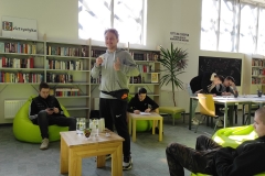Młody uśmiechnięty mężczyzna z uniesiona ręką i podniesionym do góry kciukiem, w drugiej ręce trzyma kubek. Wokół rozstawione jaskrawe siedziska, na których siedzą młodzi mężczyźni. W oddali dwie osoby pochylające się nad stołem. W tle ustawione wzdłuż ściany regały wypełnione książkami.
