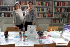Dwie uśmiechnięte kobiety stoją przy stole, na którym ułożone są kolorowe publikacje oraz gadżety promocyjne. W tle regały z książkami.