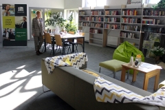 Duża przestrzeń biblioteczna.  Po lewej stronie stołu stoi kobieta. Po prawej stronie kanapa, pod scianą regały z książkami.