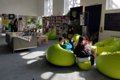 Duża przestrzeń biblioteczna. Na pierwszym planie dwie osoby siedzące na jasnozielonych pufach. W oddali stól , przy którym stoi grupa osób. Pod ścianą regały z książkami.