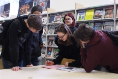 Grupa 5 licealistów pochyla się nad stołem, na którym leży różowa kartka i czasopismo „Kruszywa”. Jedna trzyma długopis. W tle regał z czasopismami.