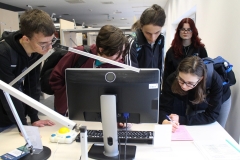 Grupa 5 licealistów pochyla się nad stołem, na którym leżą różowa kartka, klawiatura i specjalistyczna duża mysz oraz stoi monitor komputera i lampka.