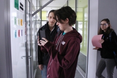 Licealista odczytuje kod QR z drzwi pokoju za pomocą telefonu. Z lewej strony stoi chłopak, z prawej dziewczyna w okularach trzyma różową kartkę.