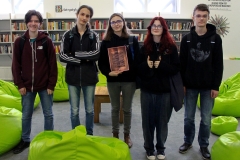 Trzech chłopców i dwie dziewczyny stoją przed regałami z książkami. Jedna z dziewczyn trzyma różową kartkę z opisem gry „Biblioteka. Złap trop”.